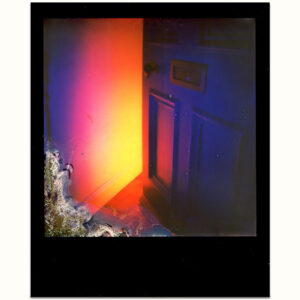 A scan of a black frame i-type Polaroid of A partially open purple door with amber yellow light radiating outwards from inside the home with a chemical burn in the lower left corner on a white background.